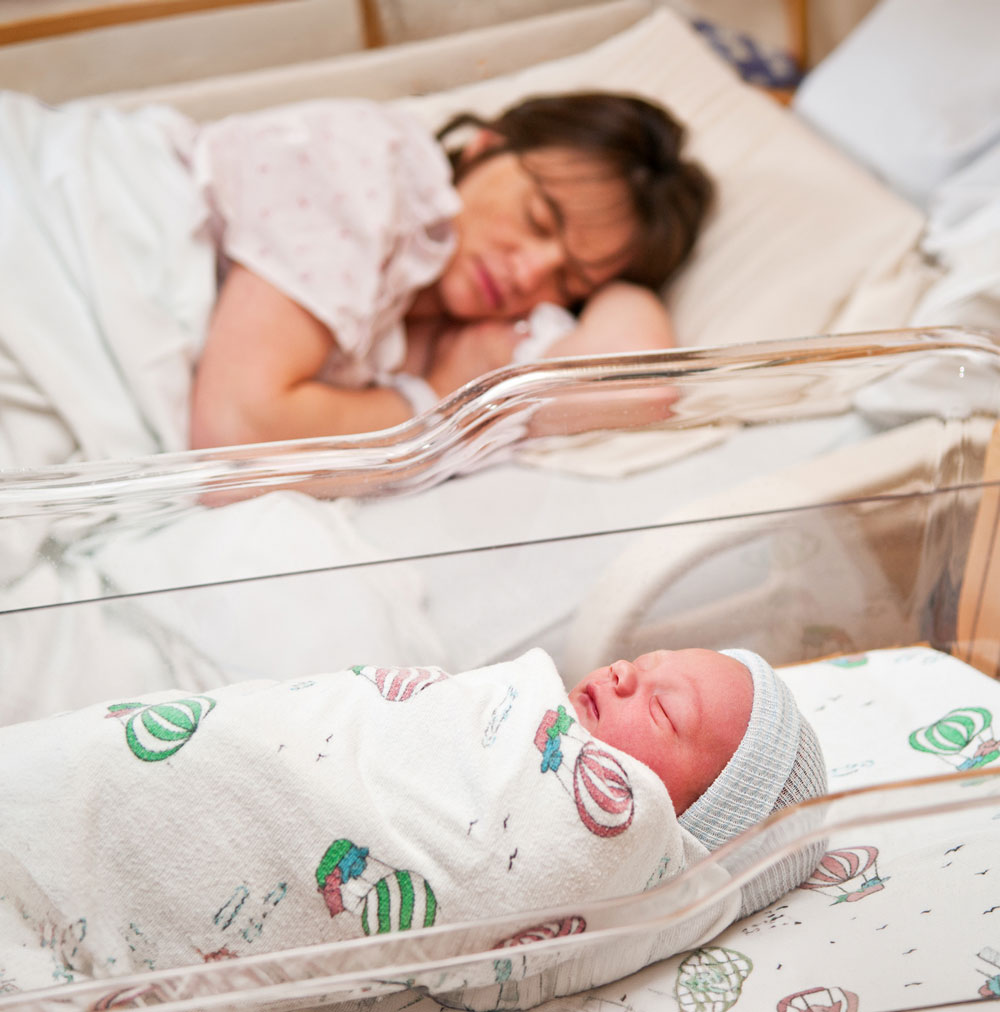 Newborn sleeping in hospital bassinet, mother resting nearby