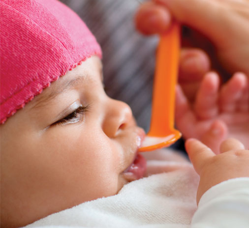 Baby eating from orange spoon, wearing pink hat.