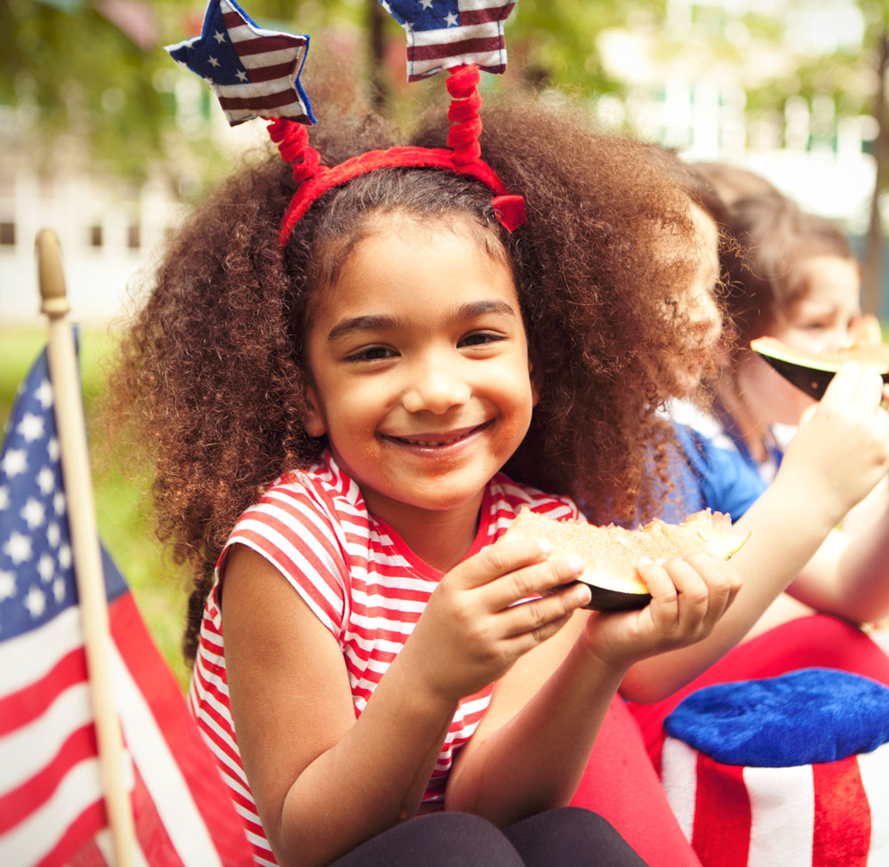 Smiling girl in patriotic headband eating watermelon by American flags