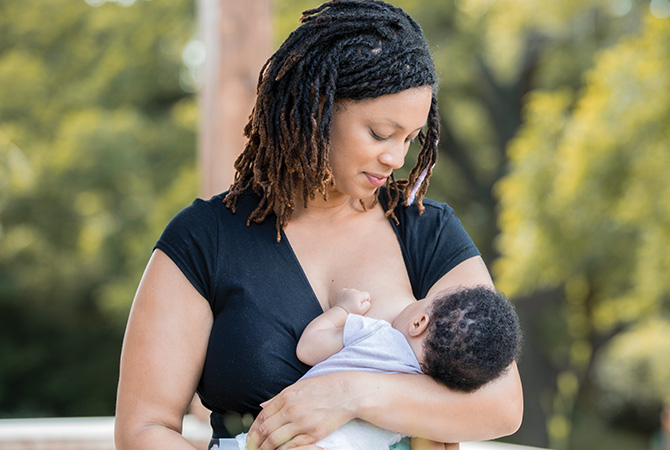 Mother breastfeeding baby outdoors, surrounded by greenery.