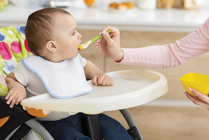 Baby being spoon-fed in highchair by caregiver.