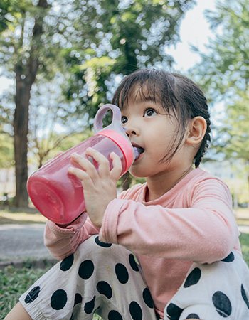 Child sitting outdoors, drinking from pink bottle